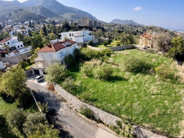 Yeşil arsa, taş duvarlarla çevrili; dağlar ve çevre evler görünüyor