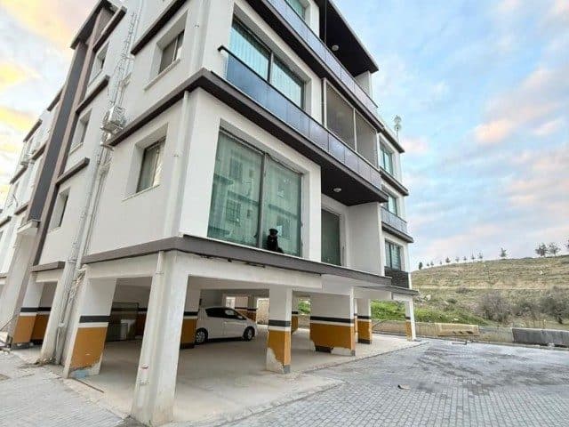 Açık otoparklı binada cam korkuluklu balkonlar görülüyor.