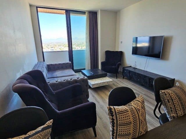 Koyu koltuklar, duvara monte televizyon ve geniş camlı balkon.