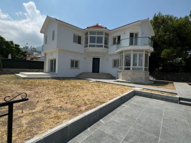 Beyaz cepheli villa, cam balkon ve taş döşeli alanla çevrili.