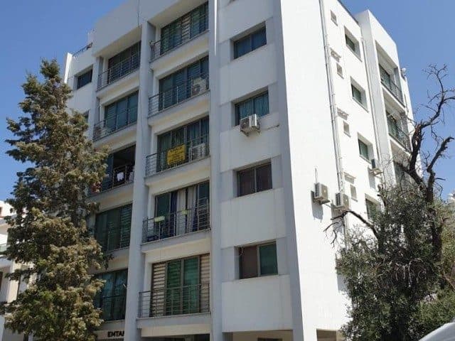 Beyaz apartman cephesi ve balkon korkulukları ağaçların arasından görünüyor.