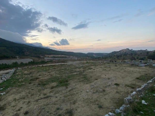Ön planda taşlı arsa, çevresinde tepeler ve bulutlu gökyüzü