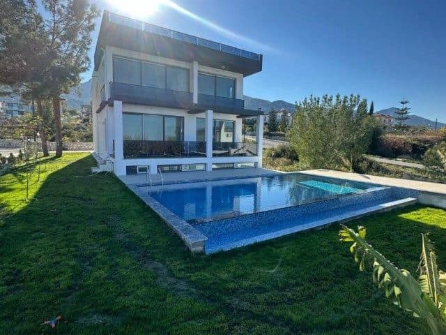 Cam balkonlu villa, dikdörtgen havuz ve çim bahçeyle görünüyor