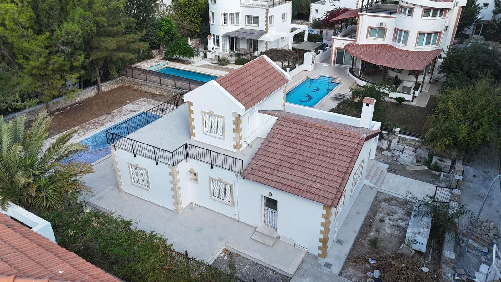 Elevated view of a white villa with red tiled roofs and a roof terrace, with nearby pools.