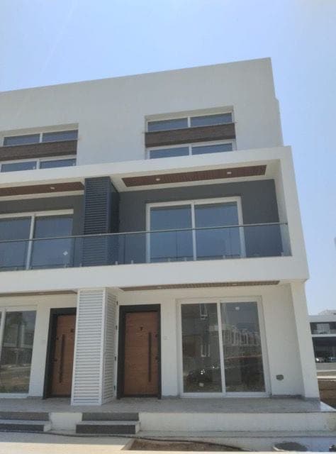 Beyaz ve gri cepheli binada ahşap giriş kapıları, geniş camlar ve cam korkuluklu balkon.
