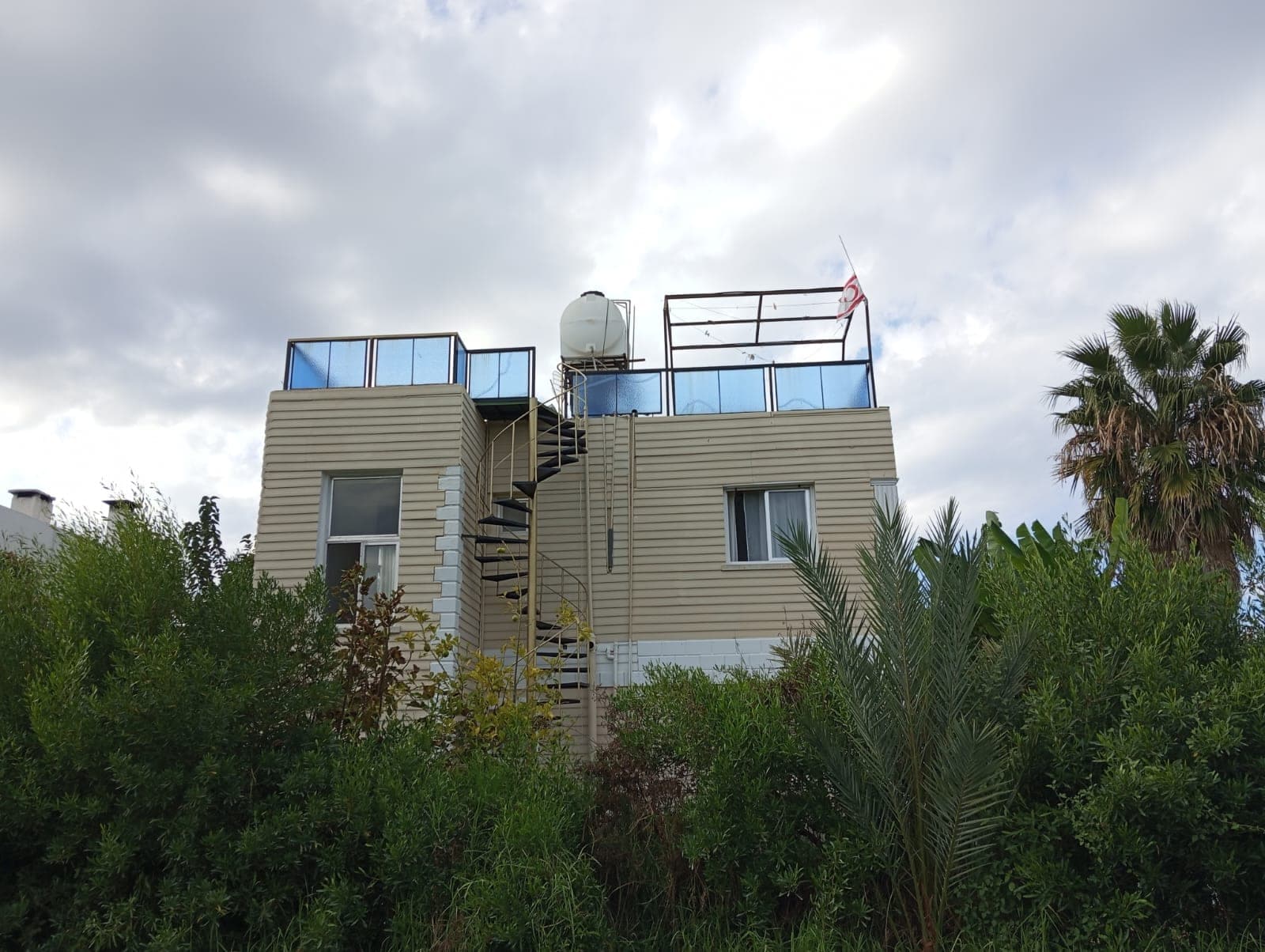 Exterior of a villa with spiral staircase, water tank on the roof, surrounded by greenery.