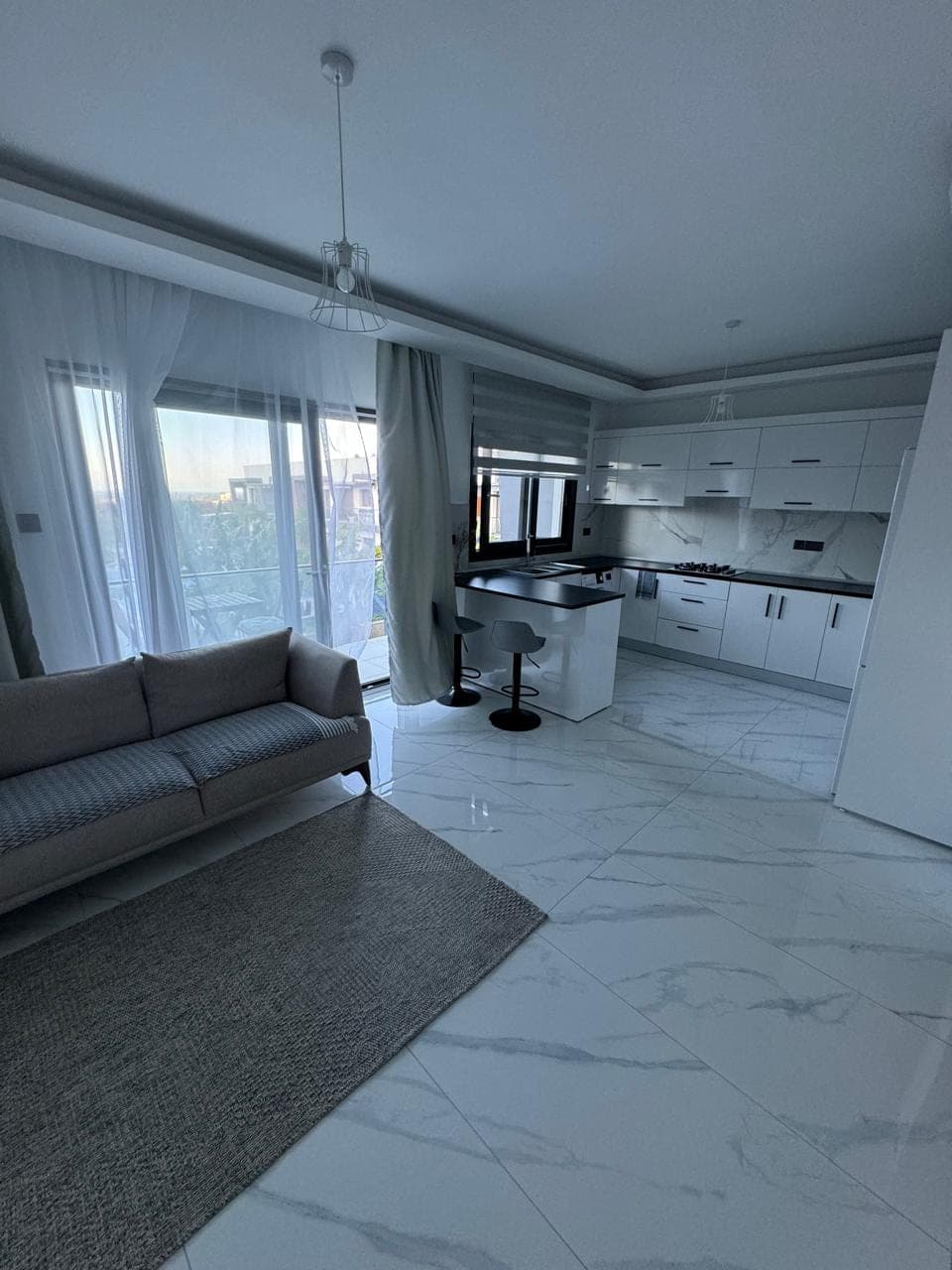 Modern open-plan living room and kitchen, opening to a balcony with large glass doors.