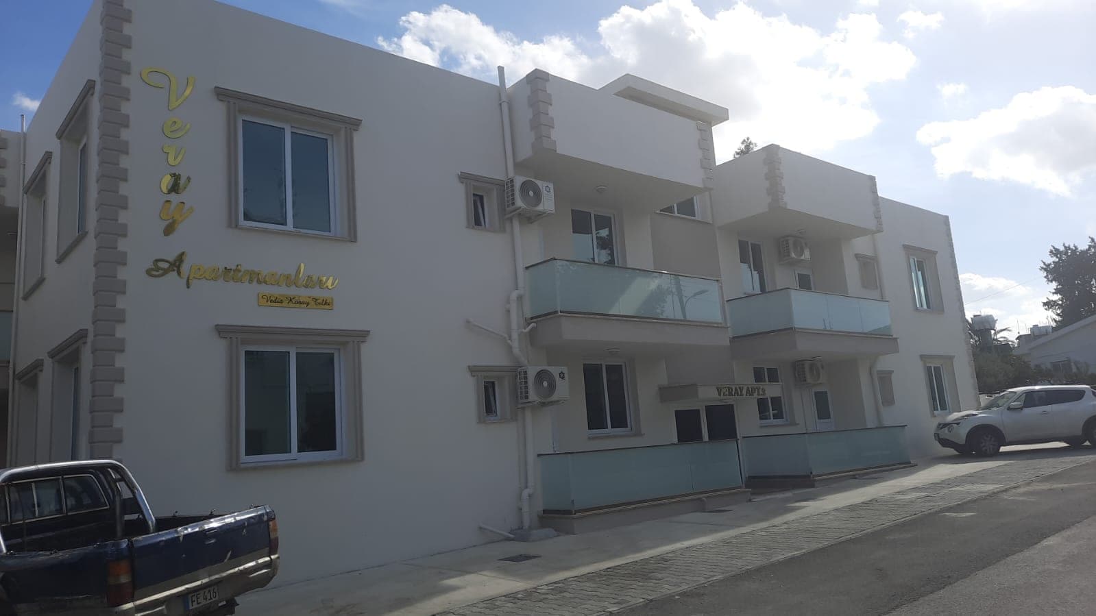 White-faced apartment building with balconies and AC units, car parked next to it.